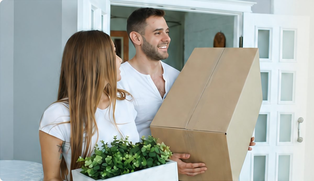 Een jong stel draagt een verhuisdoos en een plant hun nieuwe huis binnen